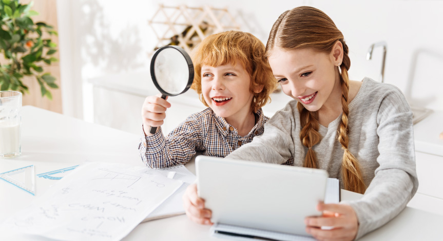 Boy looking through a magnifying glass while girl next to him looks at her computer while doing a Mad Science virtual class