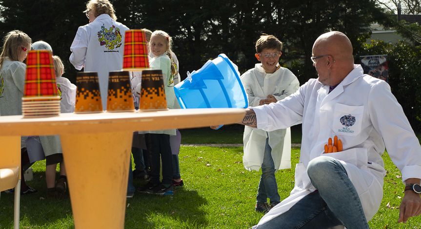 A girl in front of a large audience is holding a big yellow ball while speaking to a mad scientist holding a microphone
